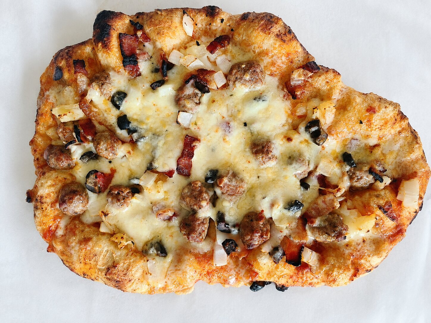 A whole rustic homemade pizza on parchment, with a puffy blistered crust, topped with sausage, bacon, onion, black olives and melted cheese.