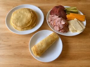 A spread of pfannkuchen on a wooden table: a stack of golden German pancakes on one plate, a plate of sliced ham, salami, cheddar, havarti, and pickles, and a rolled pfannkuchen on a small plate.