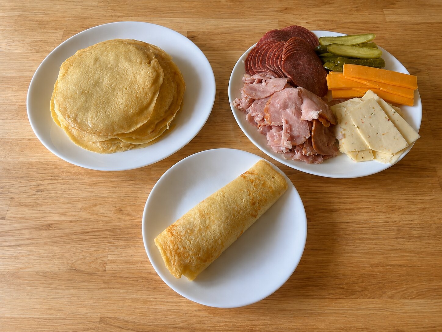A spread of pfannkuchen on a wooden table: a stack of golden German pancakes on one plate, a plate of sliced ham, salami, cheddar, havarti, and pickles, and a rolled pfannkuchen on a small plate.