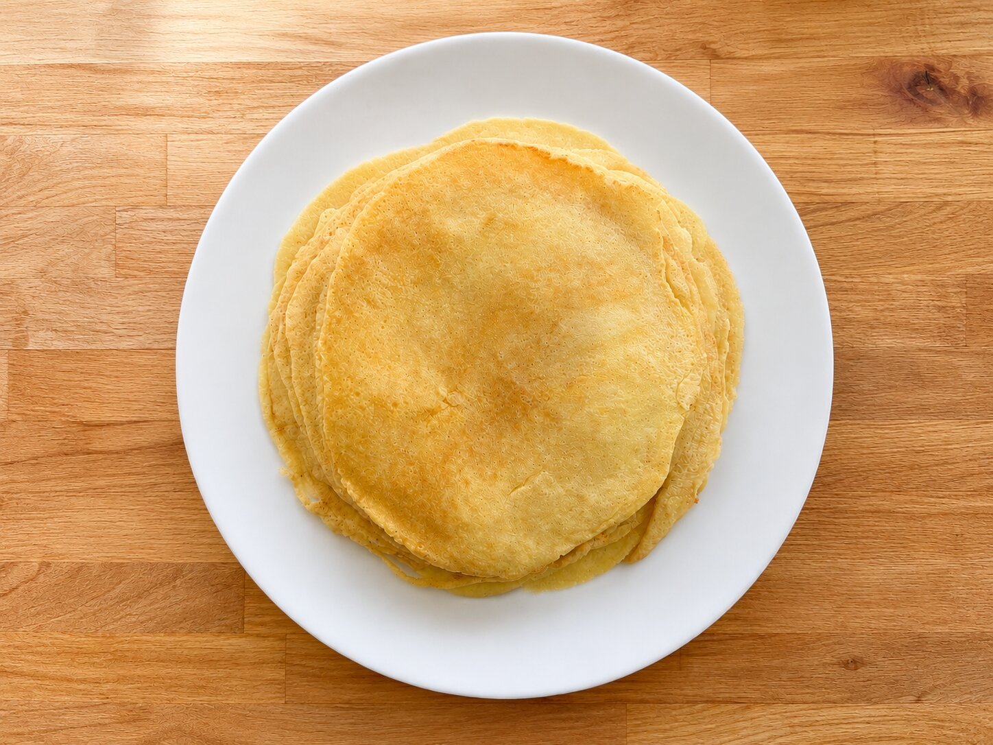 A stack of freshly made pfannkuchen seen from above on a white plate, golden and lightly browned, on a wooden table.