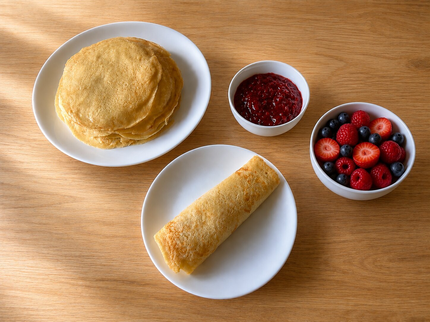 Pfannkuchen served sweet: a stack of pancakes beside a rolled pfannkuchen, a small bowl of raspberry jam, and a bowl of fresh strawberries, raspberries, and blueberries.