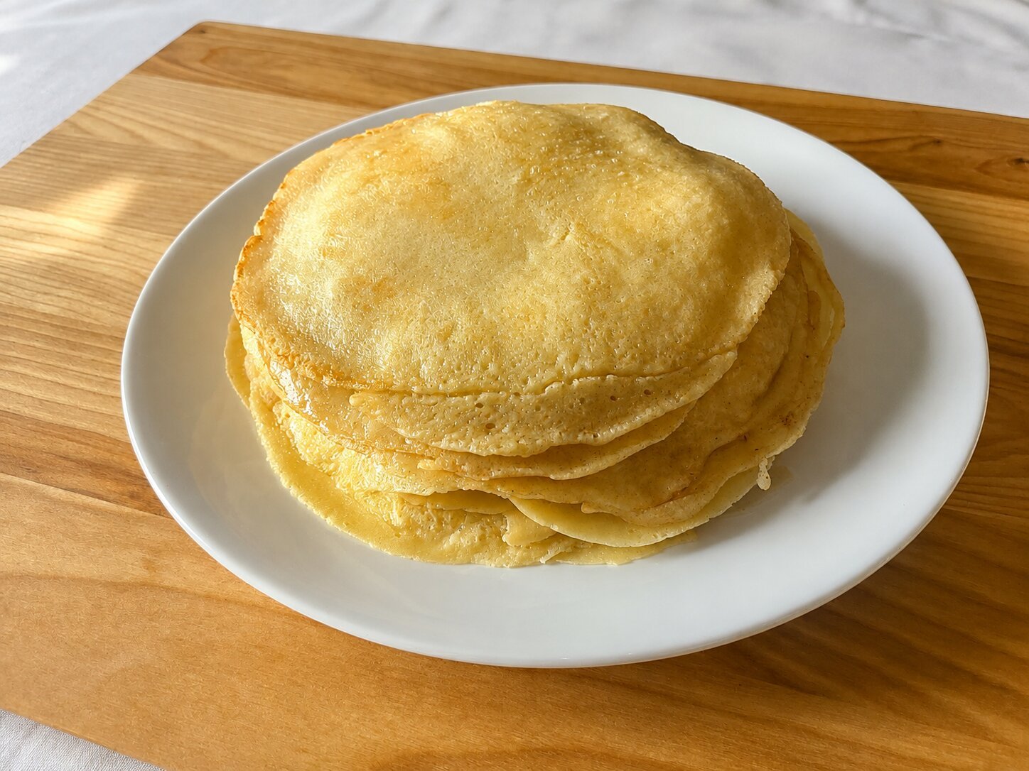 A stack of freshly made pfannkuchen on a white plate, golden and lightly browned, sitting on a wooden cutting board.